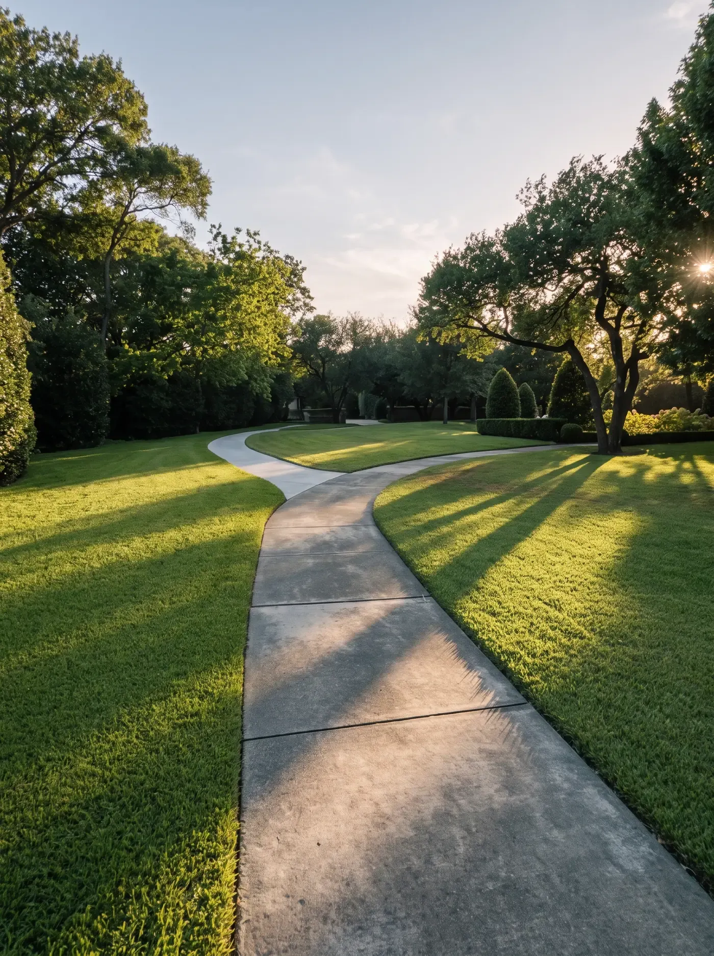 A curving concrete walk through an evenly mowed lawn under mature trees