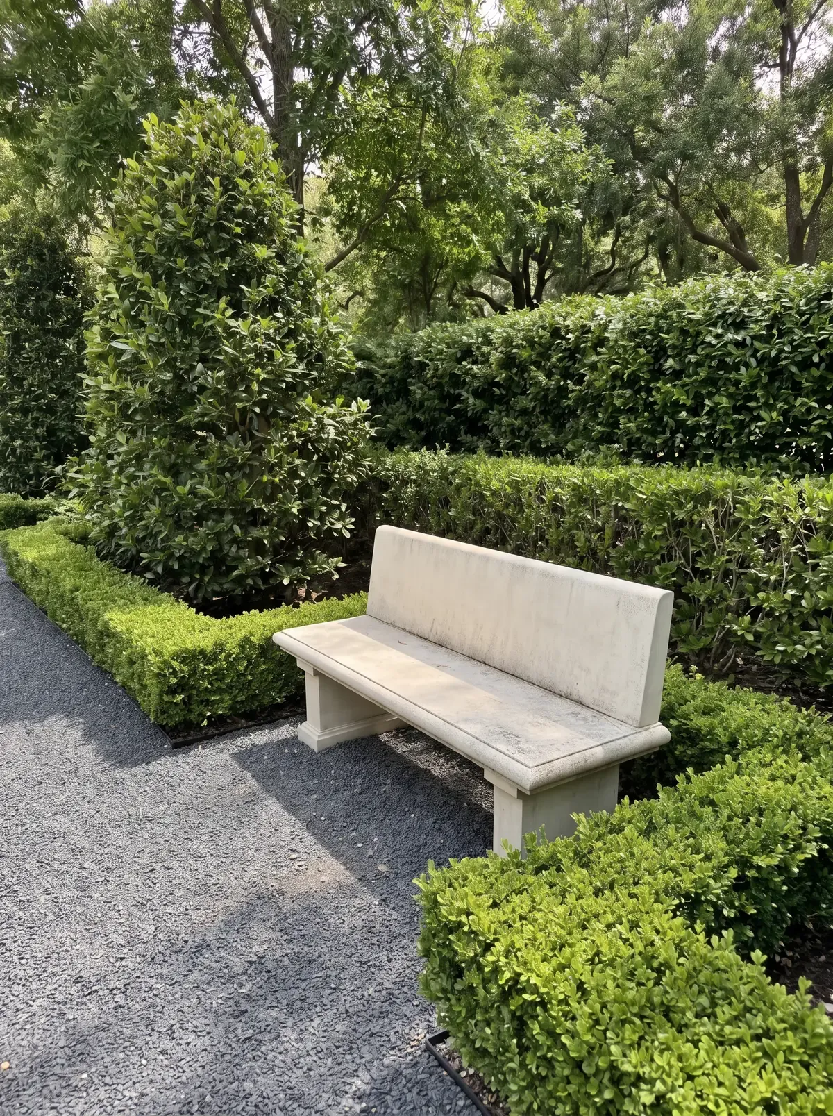 A cast-stone bench set into clipped boxwood beside a gravel path