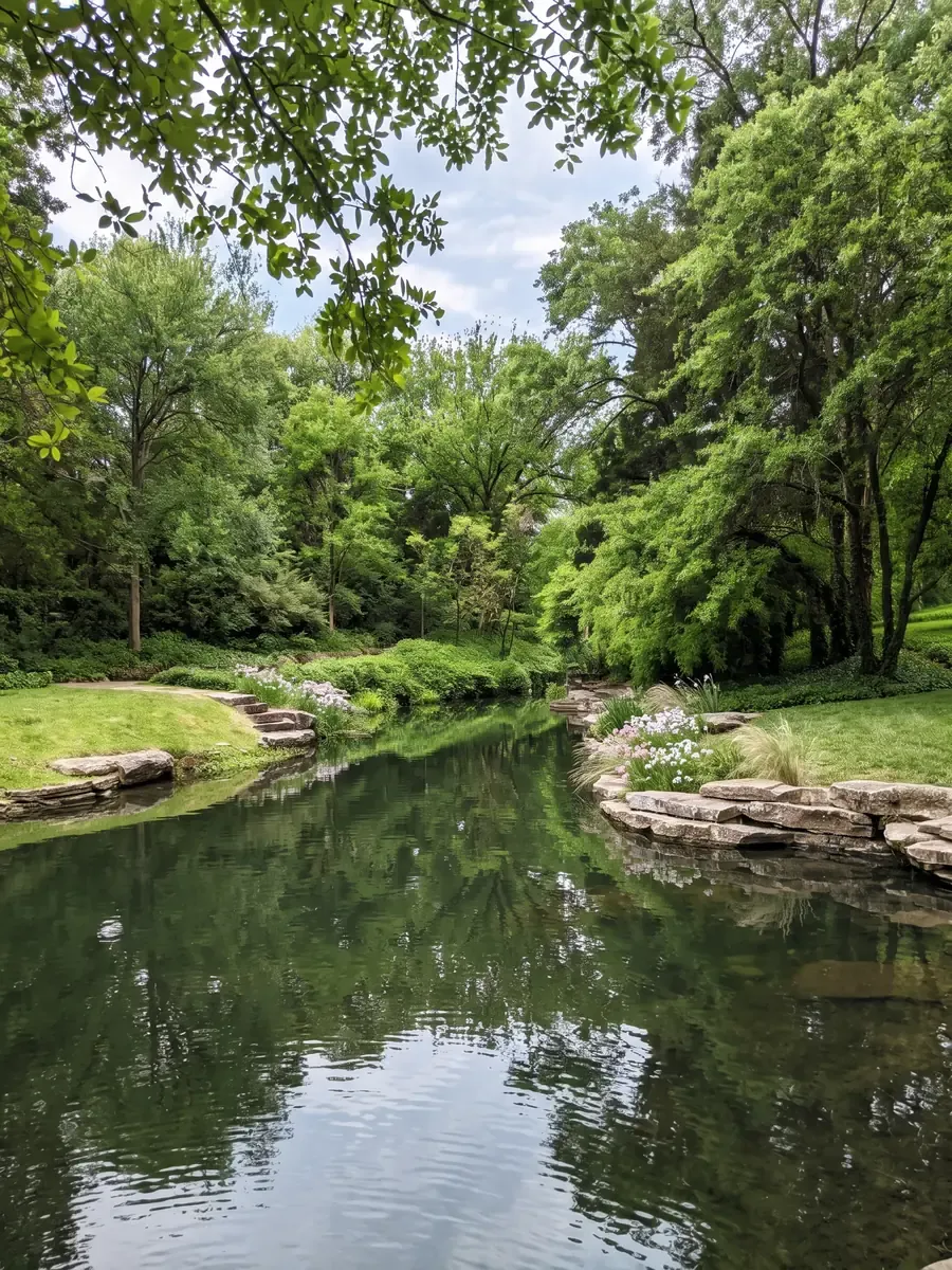 A still stream under mature trees, its banks edged with flagstone and planted with phlox and ornamental grasses, with stone steps up to the lawn